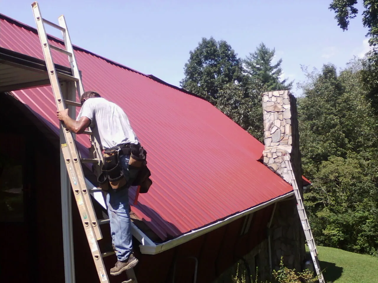 Roofer on a ladder beside a red metal roof and stone chimney — Soffit & Fascia Repair by Roofing Brothers in Paramus